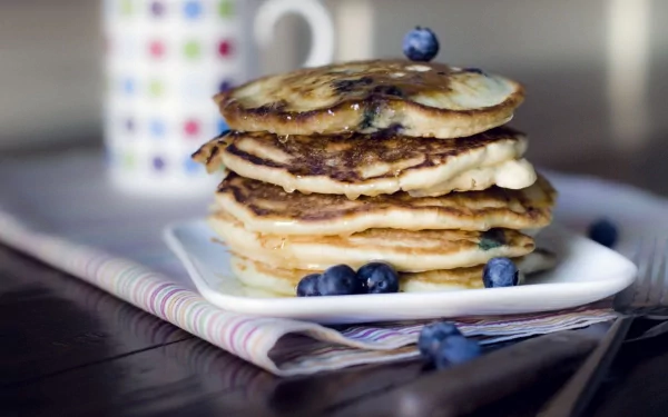 HD PC desktop wallpaper featuring a stack of golden pancakes topped with fresh blueberries on a white plate, set on a wooden table with a mug in the blurred background.