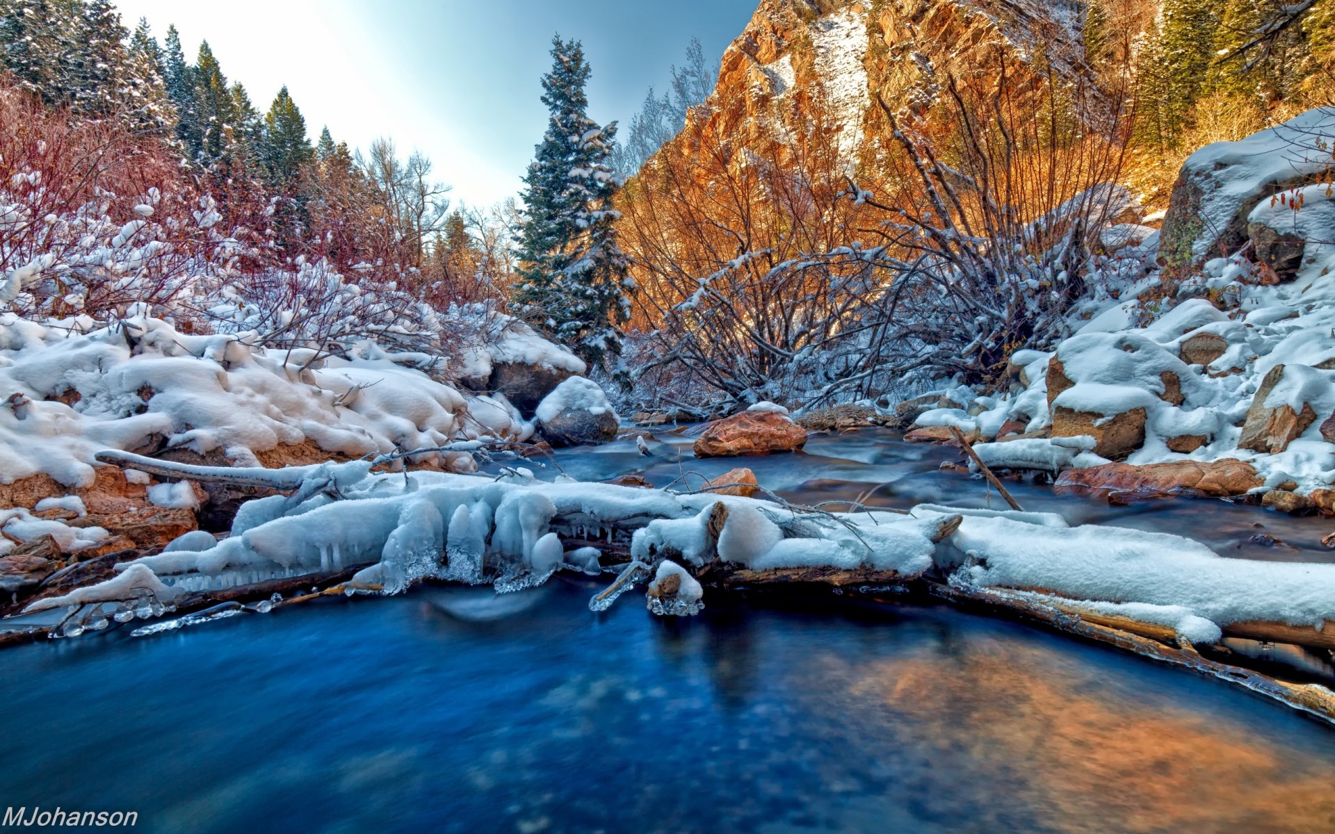 A serene winter landscape featuring a river flowing through a snowy setting, framed by trees and rocky cliffs. This HD desktop wallpaper captures the beauty of nature's tranquility.