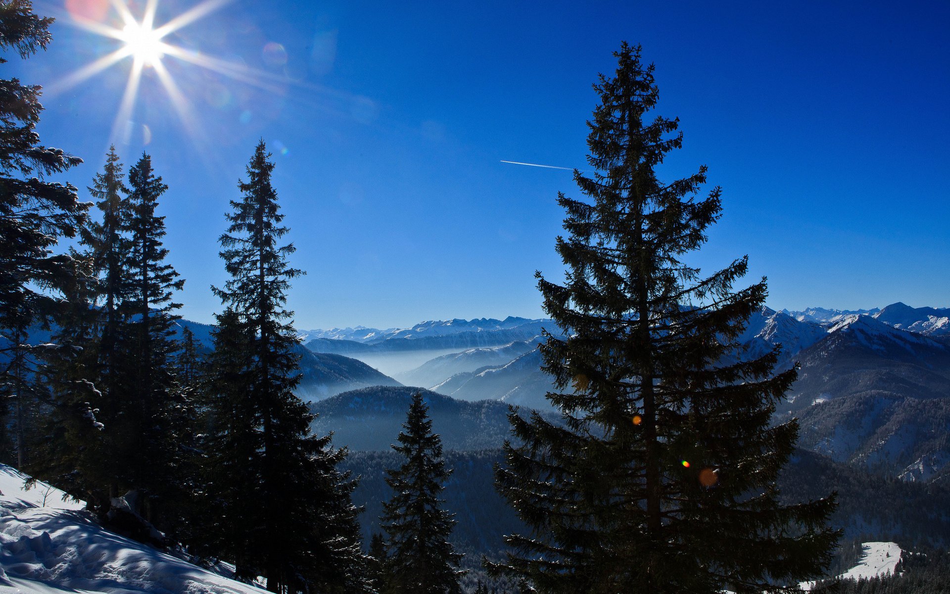HD PC desktop wallpaper featuring a bright sun over snow-covered pine trees and distant mountain ranges under a clear blue sky in a stunning natural landscape.