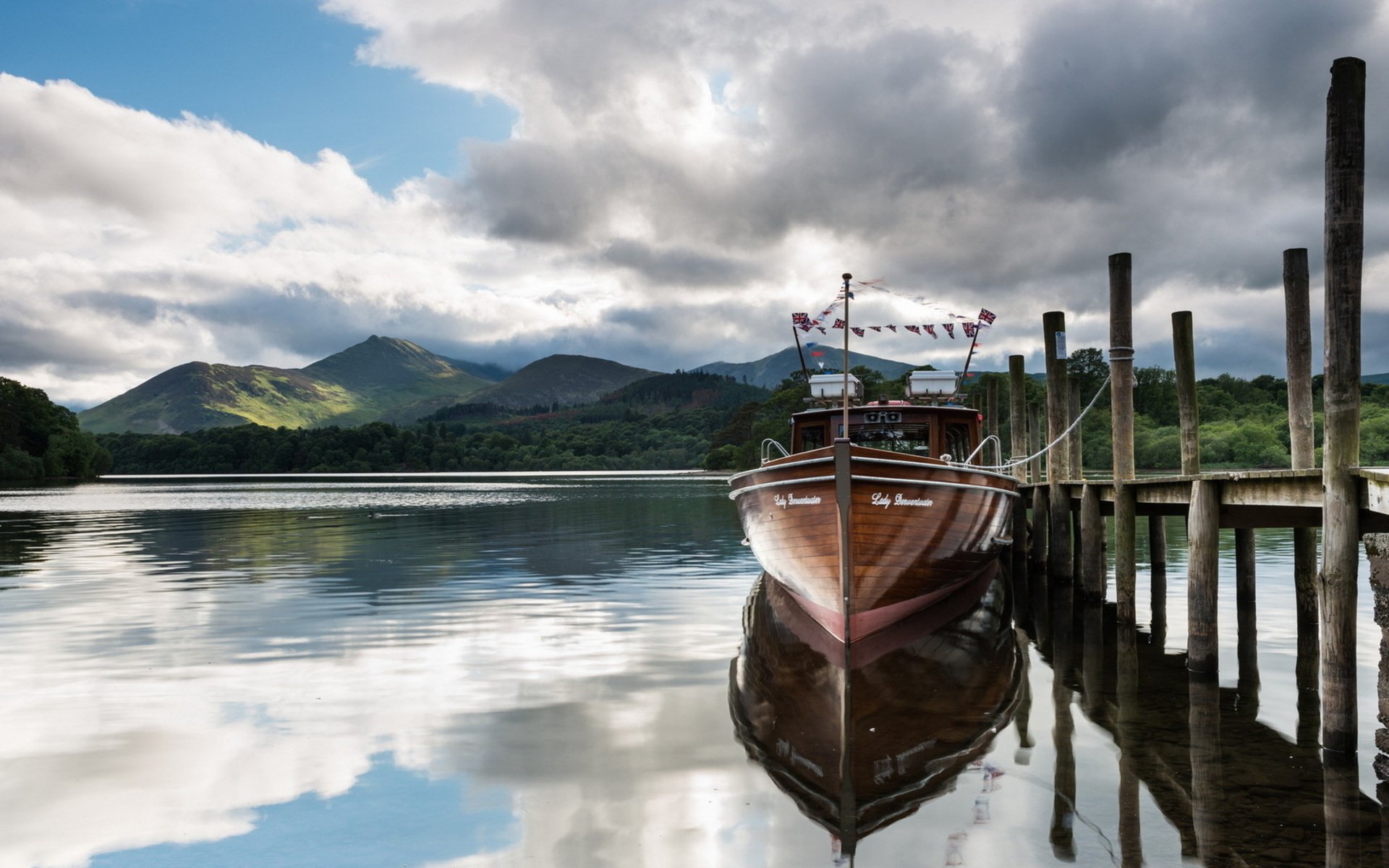 A serene scene featuring a boat docked by a wooden pier, reflecting in calm waters with mountains and a cloudy sky in the background, suitable as an HD PC desktop wallpaper.