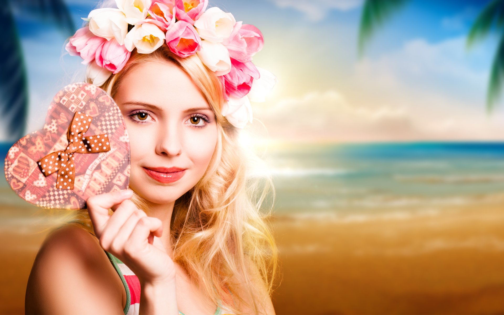 A woman with a flower crown smiles while holding a patterned heart in front of a sunset-lit beach scene, creating a vibrant HD PC desktop wallpaper background.