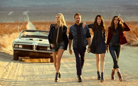 A stylish HD wallpaper featuring Paul Walker with models Thairine Garcia, Erin Heatherton, and Izabel Goulart walking confidently along a dusty road beside a classic car.