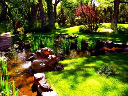 A serene garden scene with lush green grass, a small stream with rocks, tall plants, and trees in the background. The vibrant colors and tranquil setting create a peaceful HD desktop wallpaper and background.
