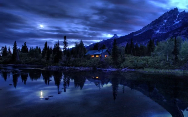 HD desktop wallpaper featuring a serene night landscape with a warmly lit house nestled among trees, near a calm reflective lake, and framed by majestic mountains under a cloudy sky.