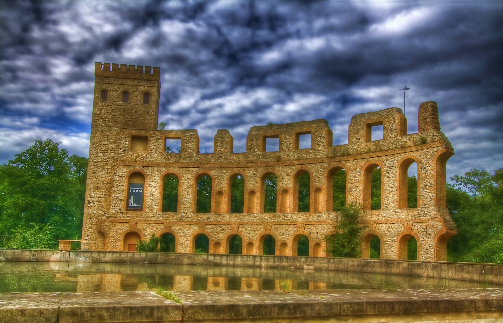 4K Ultra HD PC desktop wallpaper of a stone stadium-like ruin reflected in calm water beneath dramatic storm clouds, framed by lush green trees.
