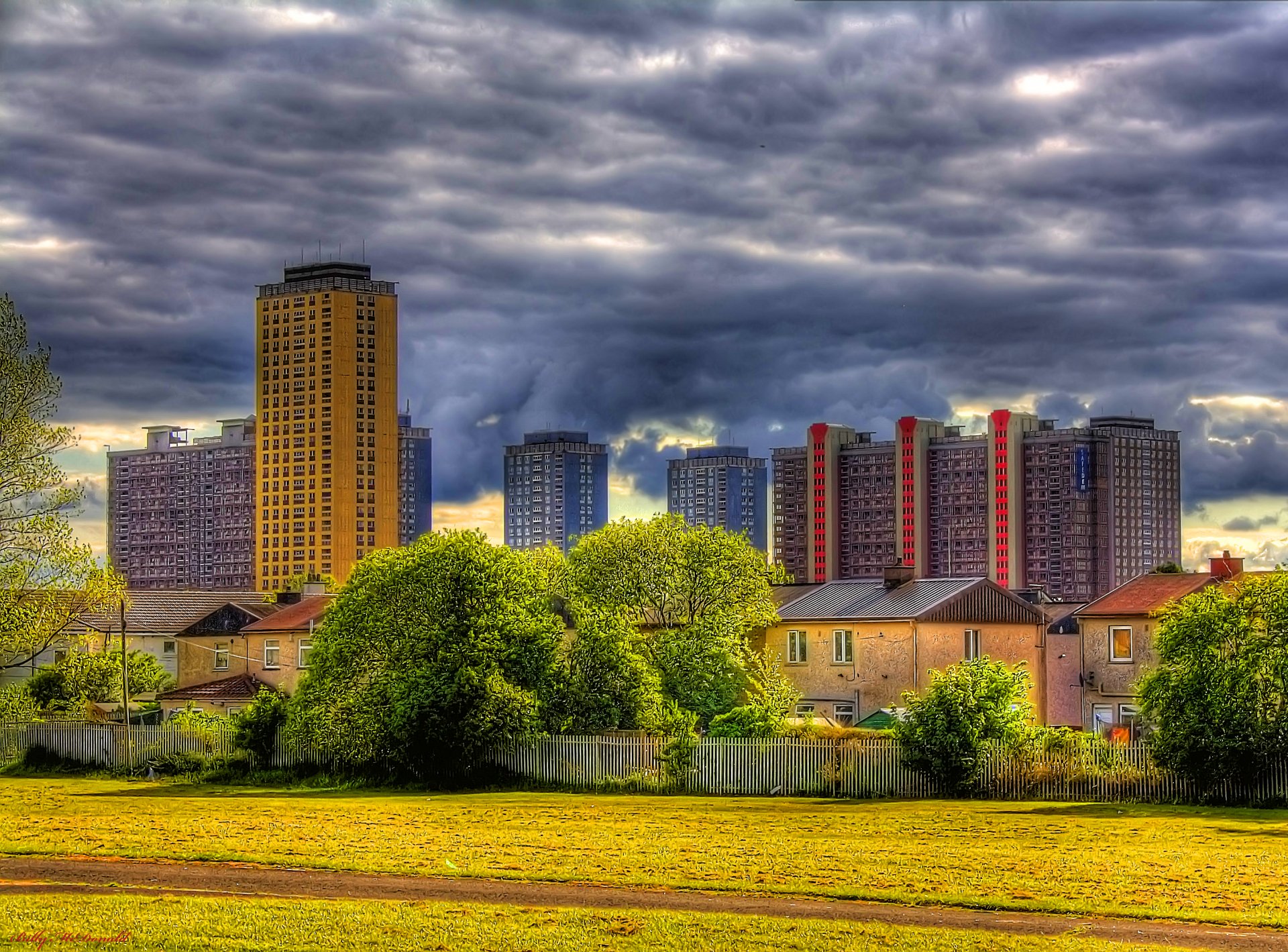 HD PC desktop wallpaper showcasing a vibrant cityscape with tall buildings under a dramatic cloudy sky, framed by lush greenery in the foreground.