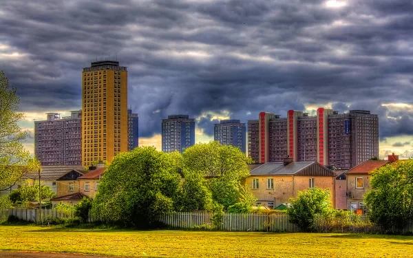 HD PC desktop wallpaper showcasing a vibrant cityscape with tall buildings under a dramatic cloudy sky, framed by lush greenery in the foreground.