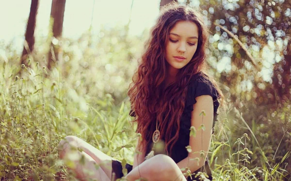 A woman with long, wavy hair sits in a sunlit field, surrounded by greenery. She gazes down softly, capturing a serene mood. This HD desktop wallpaper radiates tranquility and natural beauty.