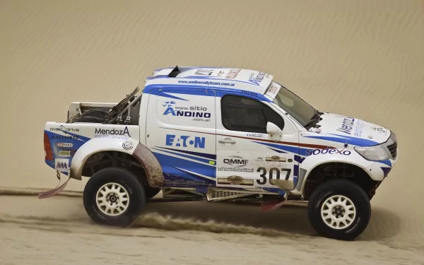 HD PC desktop wallpaper featuring a Toyota Hilux off-road racing vehicle covered in sponsor decals, driving through a sandy terrain.