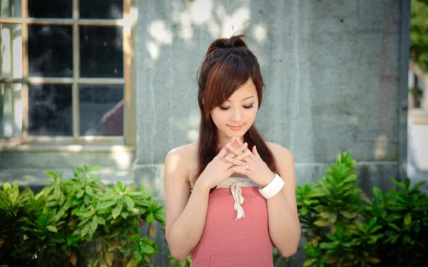HD desktop wallpaper featuring Mikako Zhang Kaijie, a woman in a coral dress standing in front of a window and greenery, with her hands clasped and looking down thoughtfully.