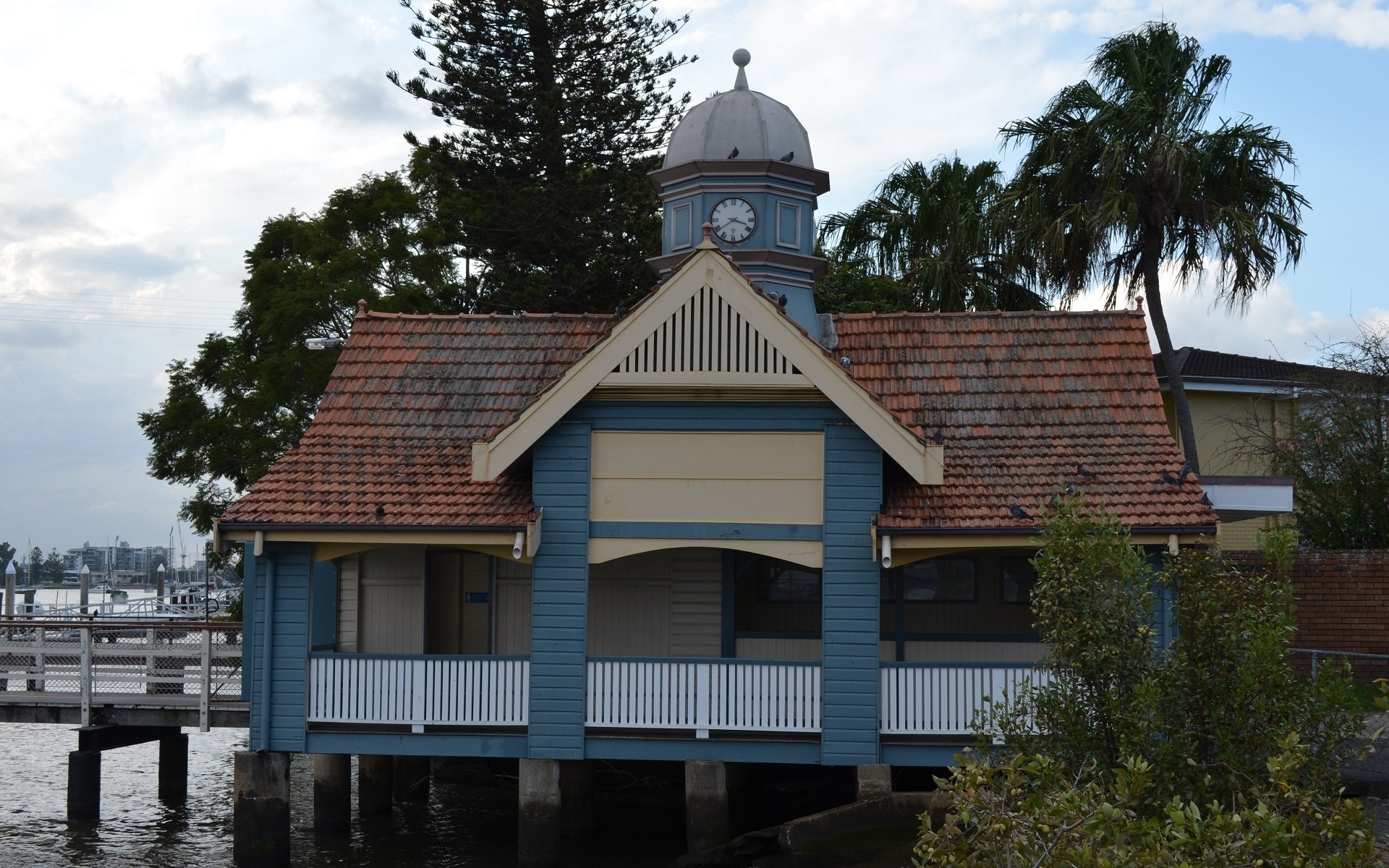 HD desktop wallpaper of a man-made building by the water in Brisbane, Australia, featuring a clock tower and surrounded by trees under a cloudy sky.