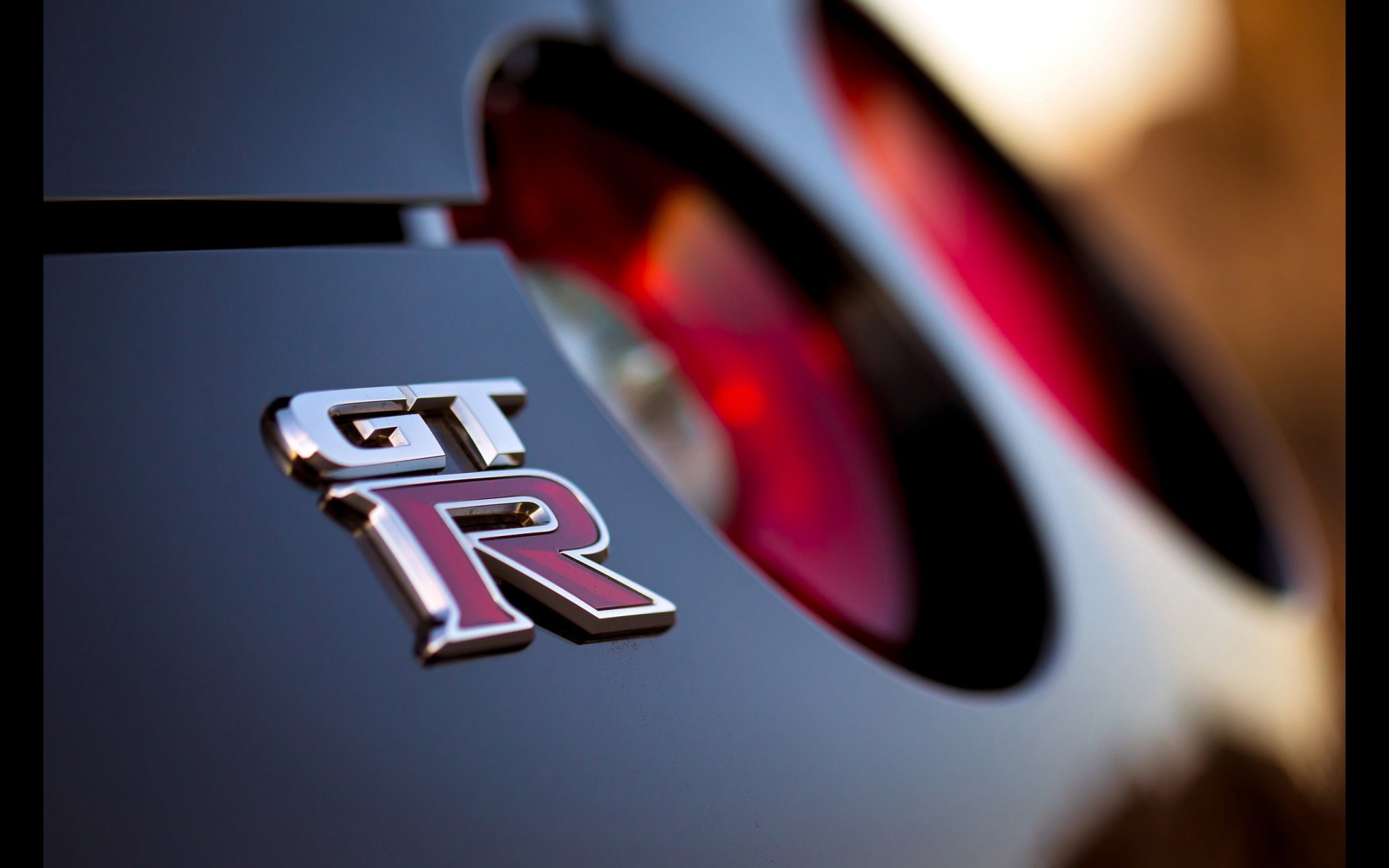 Close-up of Nissan GT-R vehicle rear badge and taillight on glossy body with blurred background — 2K Quad HD PC desktop wallpaper and background.