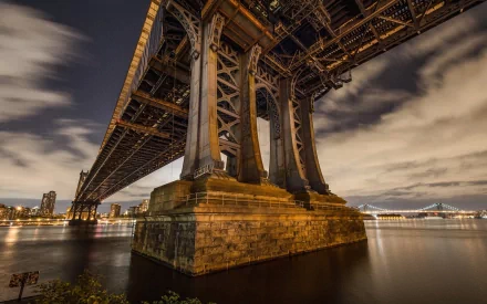 A captivating view of the Manhattan Bridge, showcasing its towering supports and intricate metalwork, set against a serene waterfront at night. An impressive HD desktop wallpaper.
