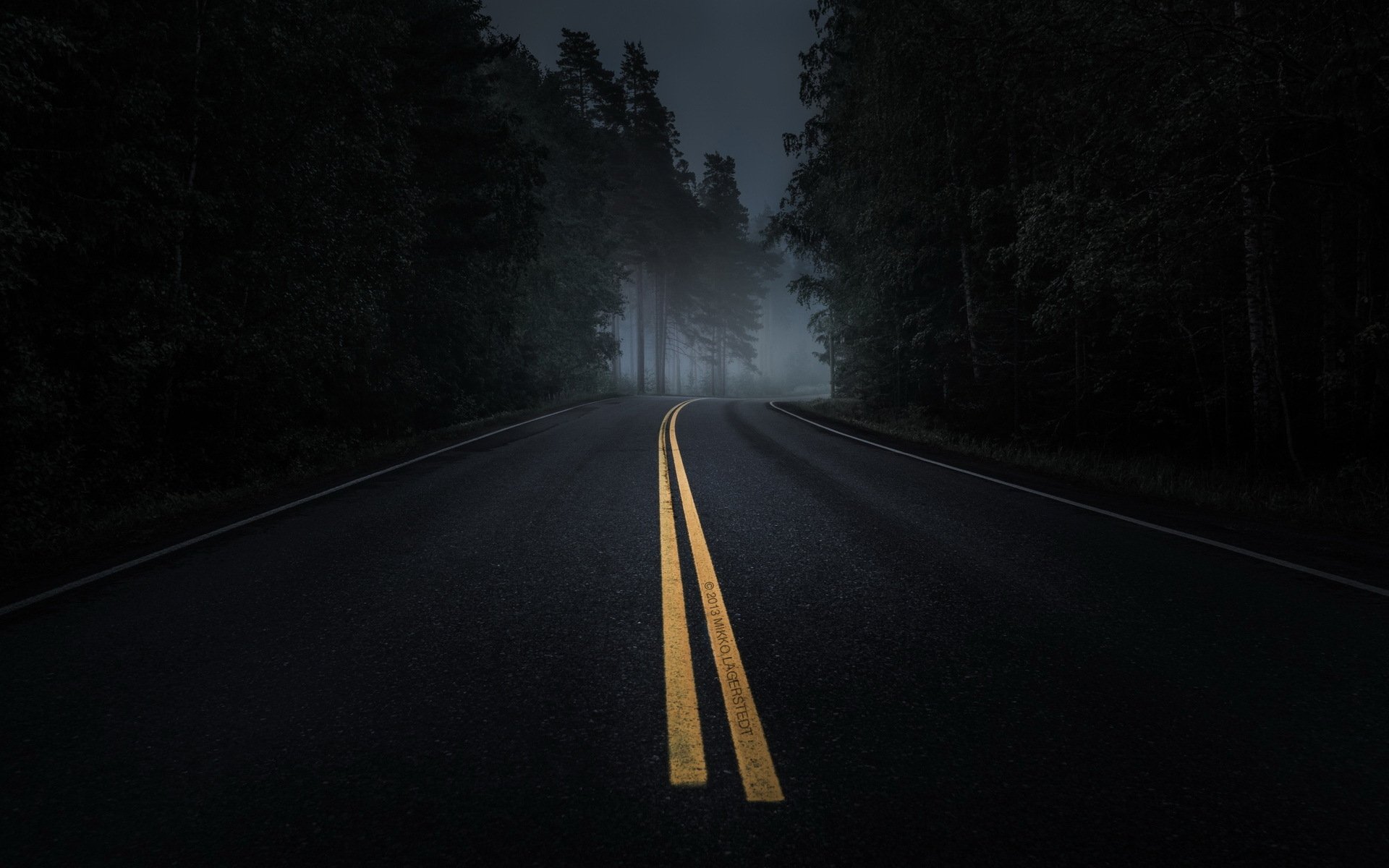 A foggy, man-made road stretches into the distance, flanked by shadowy trees, creating a mysterious and atmospheric scene for a HD desktop wallpaper.