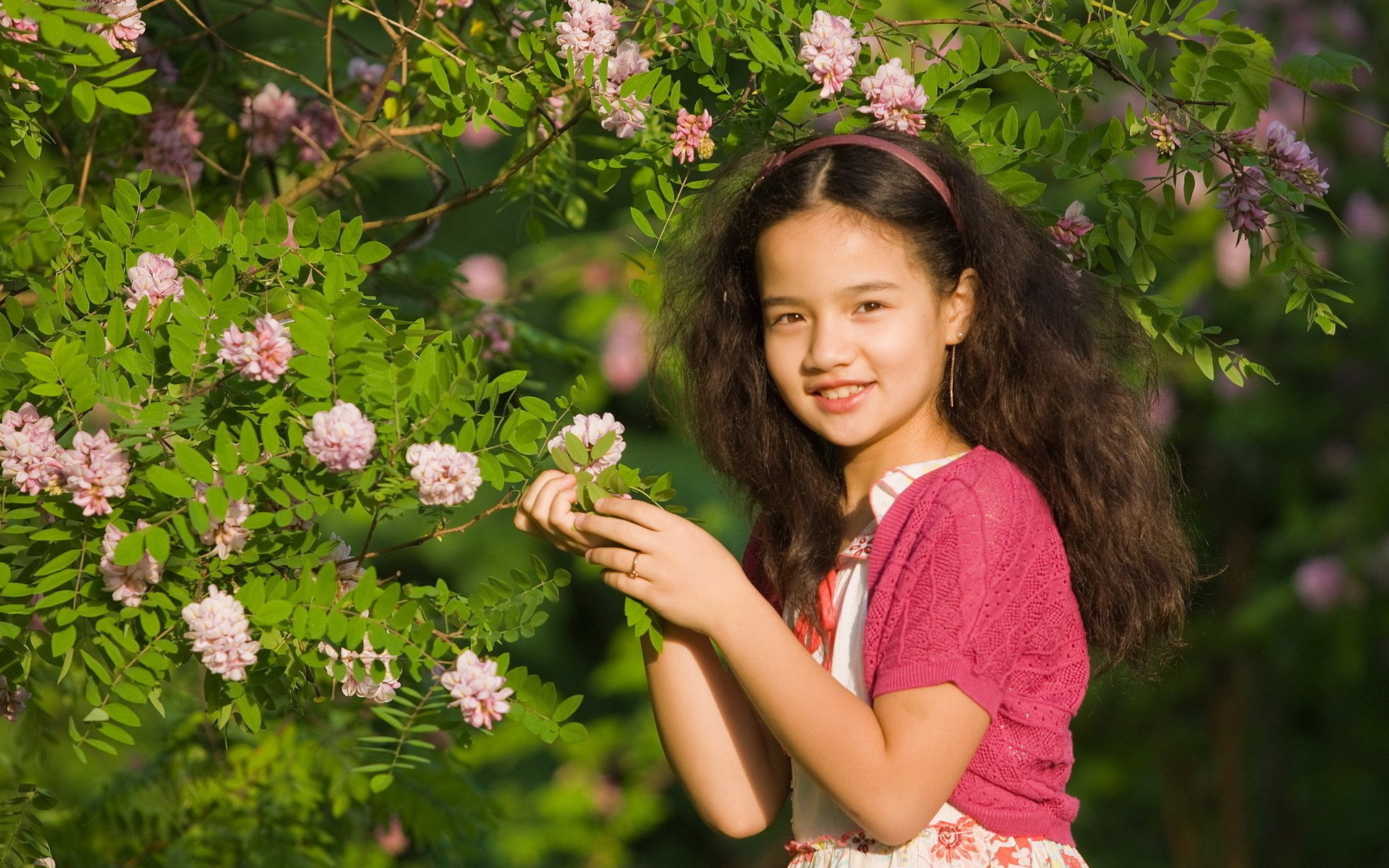 HD desktop wallpaper featuring a smiling child with long hair, wearing a pink outfit, surrounded by flowering greenery in bright natural light.