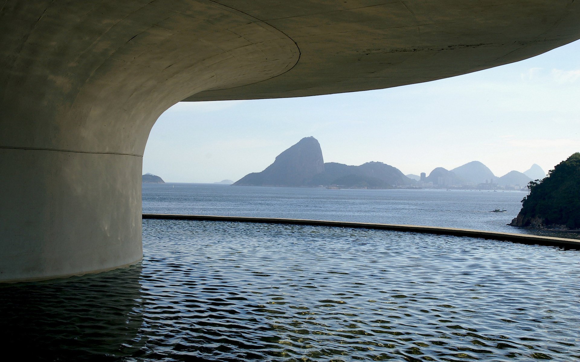 HD PC desktop wallpaper showing a serene water view framed by a curved concrete structure, with distant mountains and a calm sea under a clear sky.