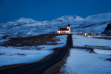 A serene church stands illuminated against a twilight backdrop, surrounded by snow-covered mountains and an empty road, creating a tranquil religious scene in this HD desktop wallpaper.