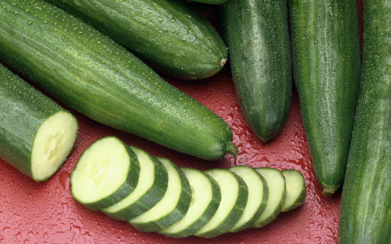 HD PC desktop wallpaper featuring fresh cucumbers, some whole and some sliced, arranged on a red surface, highlighting vibrant green tones and food textures.