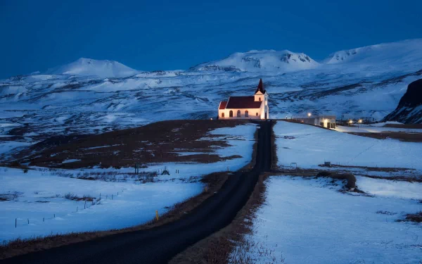 A serene church stands illuminated against a twilight backdrop, surrounded by snow-covered mountains and an empty road, creating a tranquil religious scene in this HD desktop wallpaper.