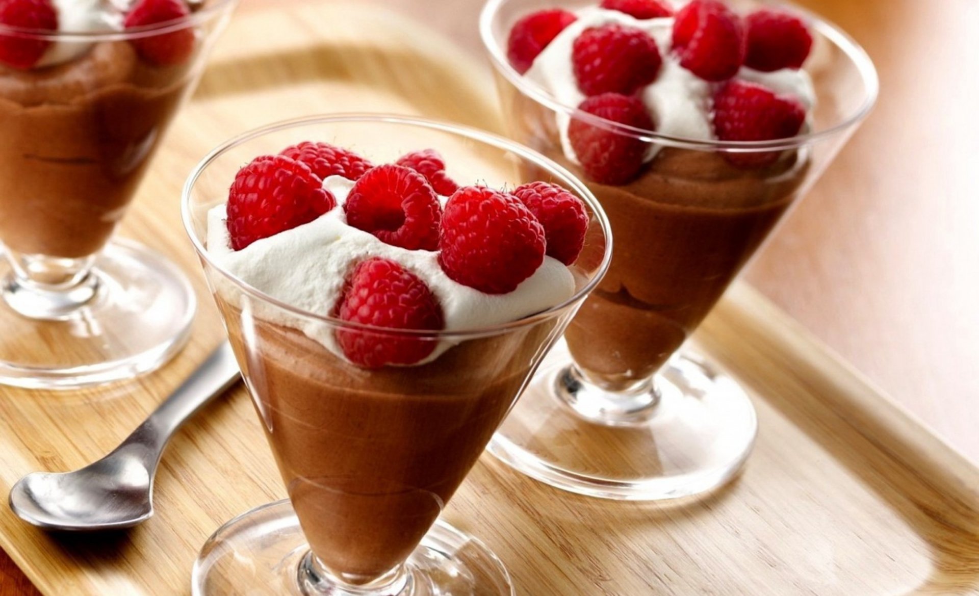 Decadent chocolate mousse topped with whipped cream and fresh raspberries, presented in elegant glass cups on a wooden tray. A delightful dessert wallpaper for food enthusiasts.
