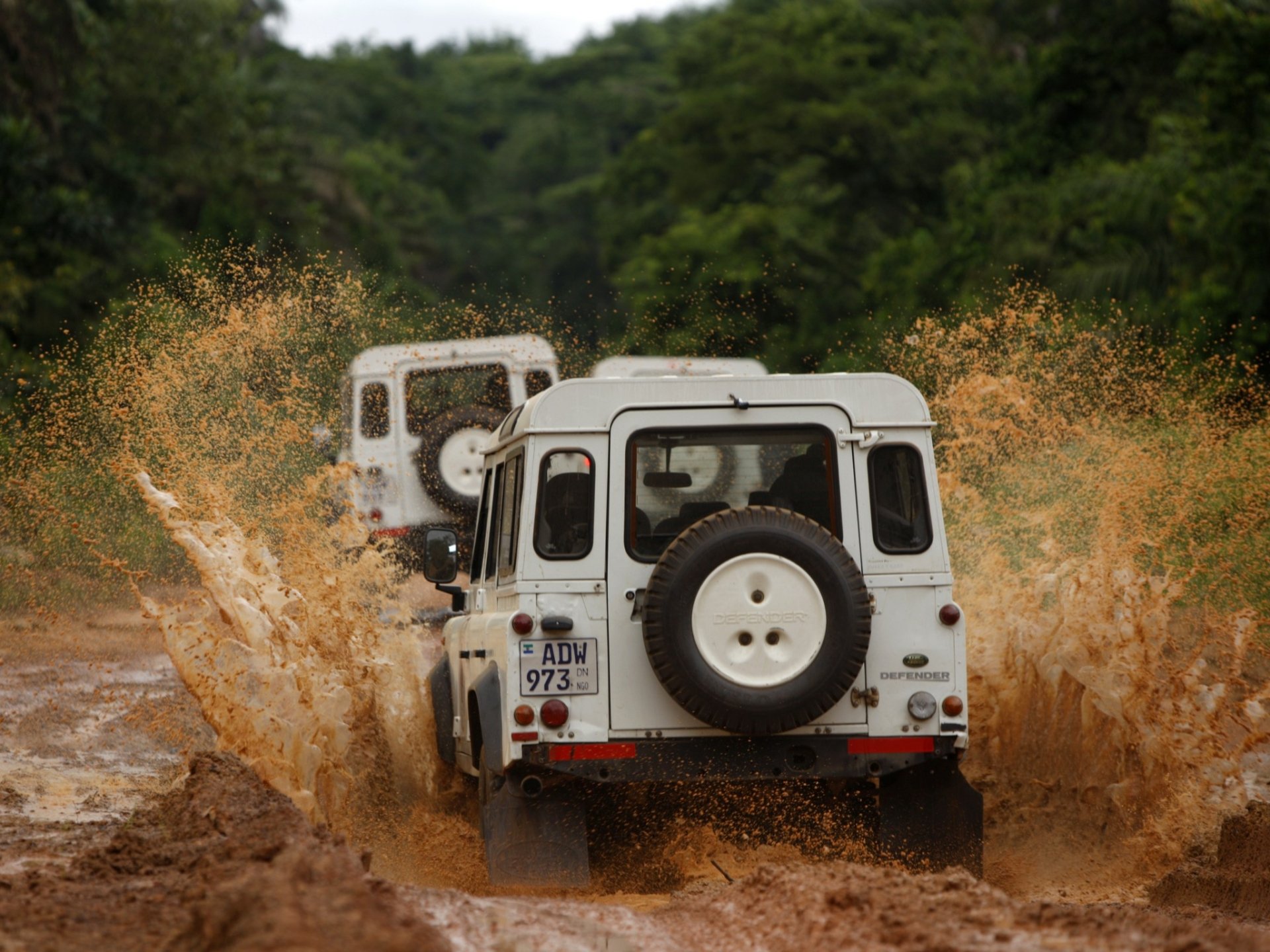 HD desktop wallpaper featuring a white Land Rover Defender driving through muddy terrain with water splashing around, set against a green forest backdrop.