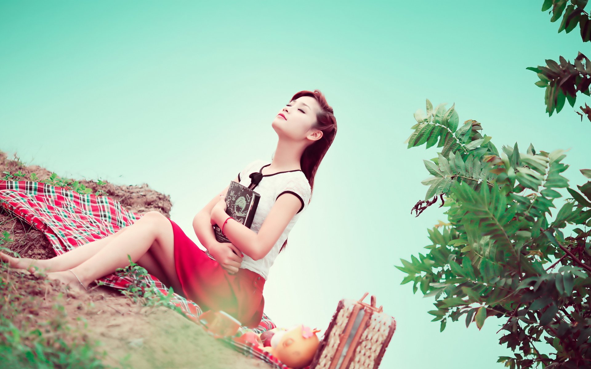 Asian woman sitting on a blanket in a natural setting, holding a book, and looking serene, HD wallpaper background.