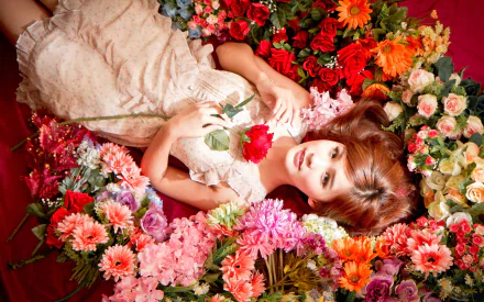 Asian woman with brown hair and eyes, wearing a white dress, lying amongst vibrant flowers while holding a rose, presented as a HD desktop wallpaper.