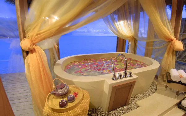 A serene ocean-view holiday spa with a man-made bathtub filled with flower petals, draped curtains, and spa essentials in an HD PC desktop wallpaper setting.