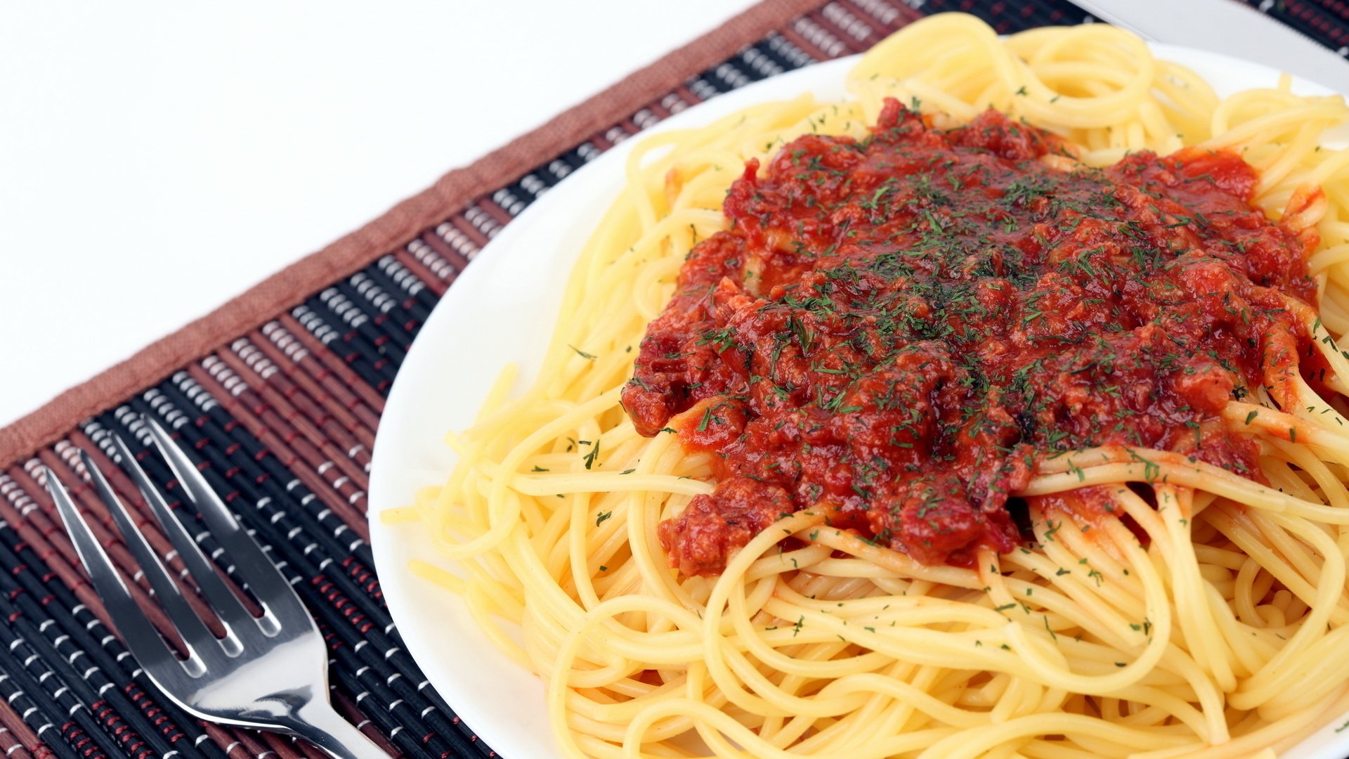 HD desktop wallpaper featuring a plate of pasta topped with rich red tomato sauce and garnished with herbs, set beside silver cutlery on a woven placemat.