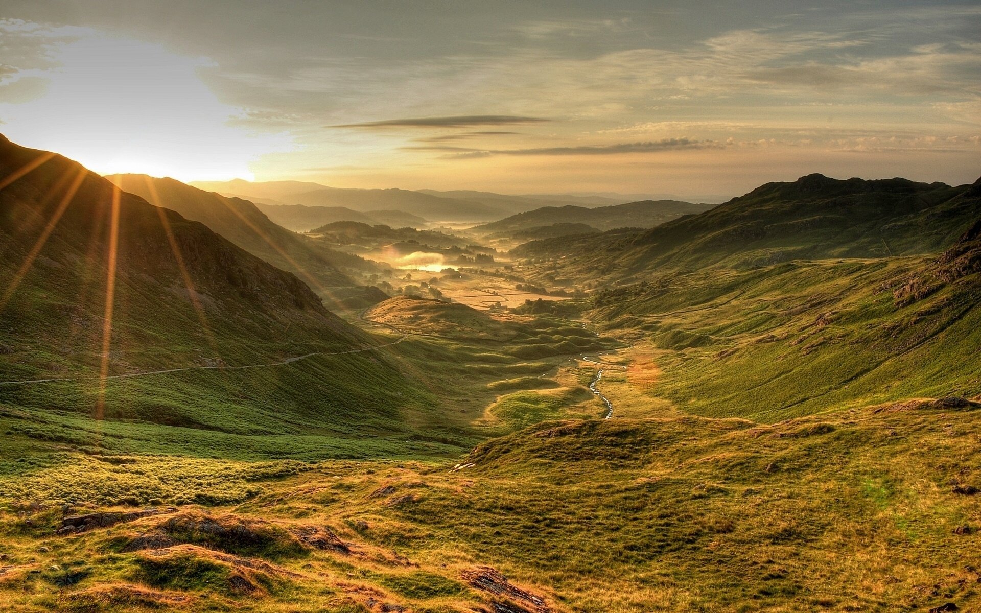 HD PC desktop wallpaper showcasing a serene nature landscape with sun rays breaking over rolling green hills and a winding valley below.