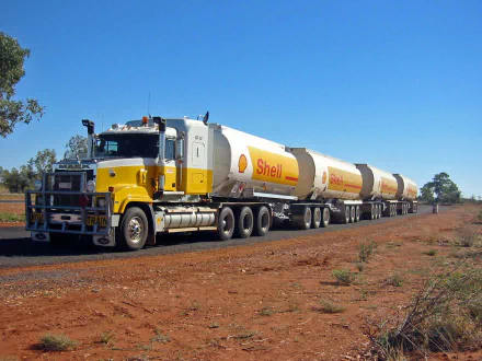 HD desktop wallpaper featuring a yellow Mack Trucks road train with Shell tankers driving on a dusty rural road under a clear blue sky.