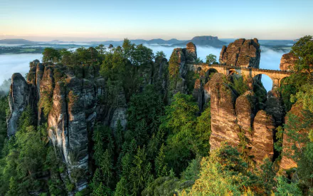 A breathtaking view of the Elbe Sandstone Mountains featuring the Bastei Bridge, surrounded by lush forests and stunning mountain landscapes under a clear sky.