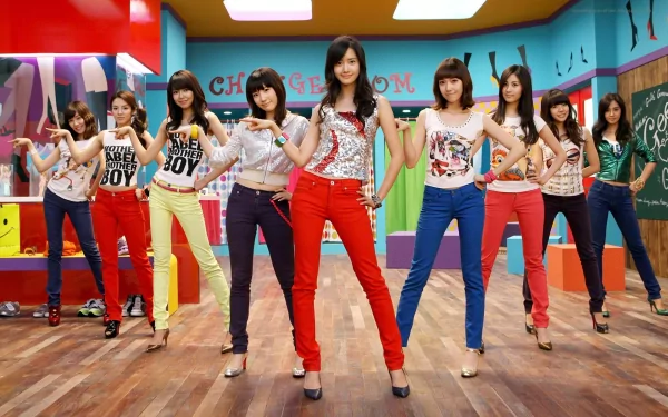 HD PC wallpaper showing nine young women in colorful pants and graphic tees striking poses in a bright music-studio set, energetic pop girl-group formation.