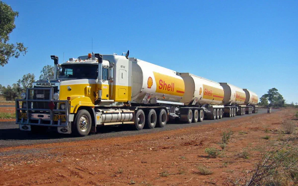 HD desktop wallpaper featuring a yellow Mack Trucks road train with Shell tankers driving on a dusty rural road under a clear blue sky.