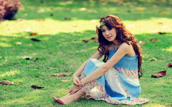 Asian woman with long brunette hair and a wreath, wearing a blue dress, sitting barefoot on grass among scattered leaves, smiling. HD desktop wallpaper.
