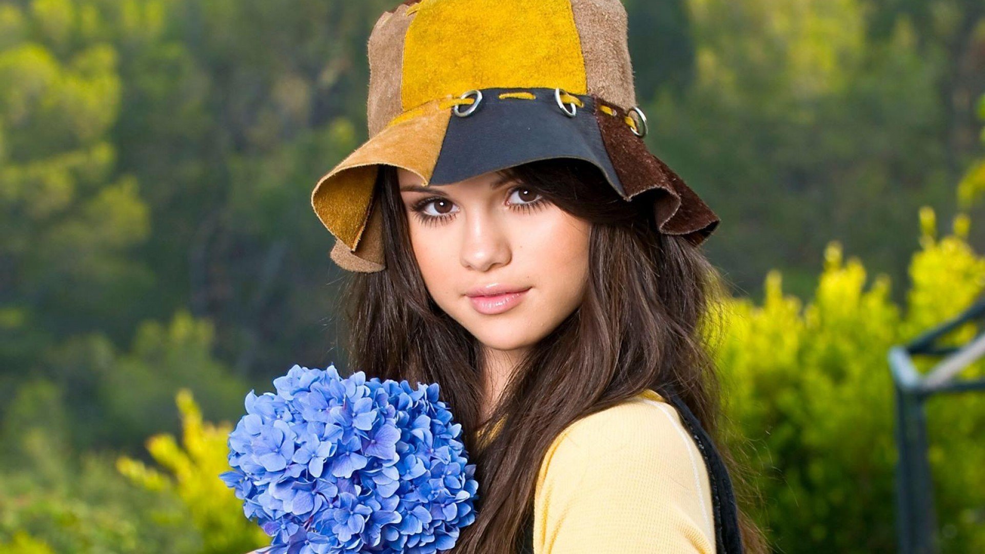 A vibrant HD wallpaper featuring Selena Gomez wearing a stylish hat and holding a blue flower, set against a lush green background, capturing her youthful charm and music-inspired essence.