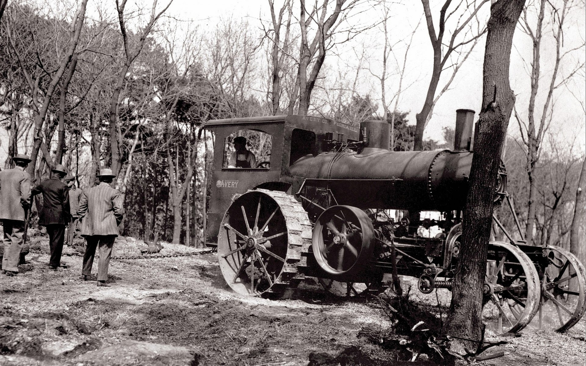 HD desktop wallpaper featuring a vintage tractor vehicle parked on a dirt path surrounded by leafless trees and two people in a forest setting.