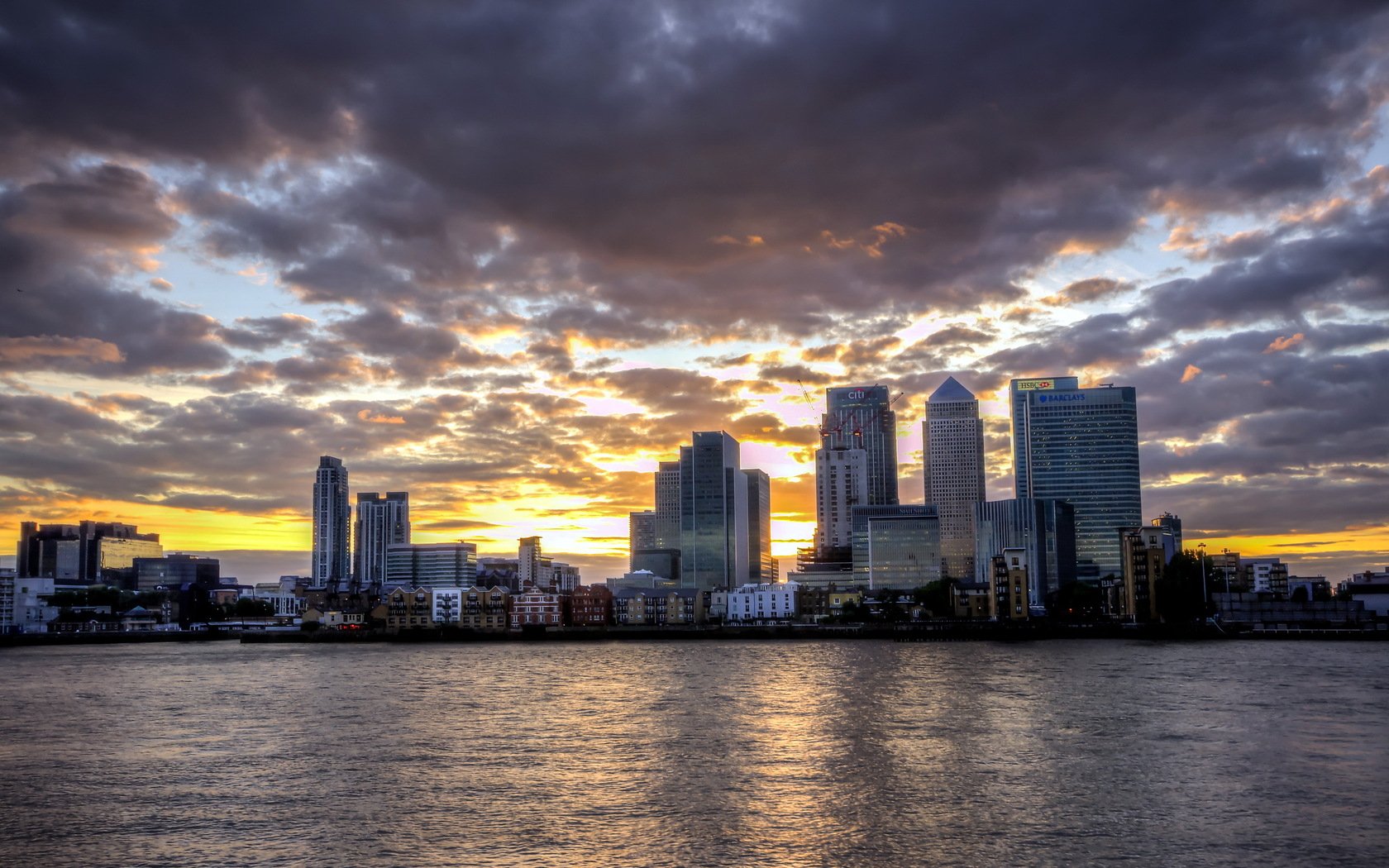 HD PC desktop wallpaper: London man-made skyline of Canary Wharf towers at sunset, dramatic clouds and river reflections.