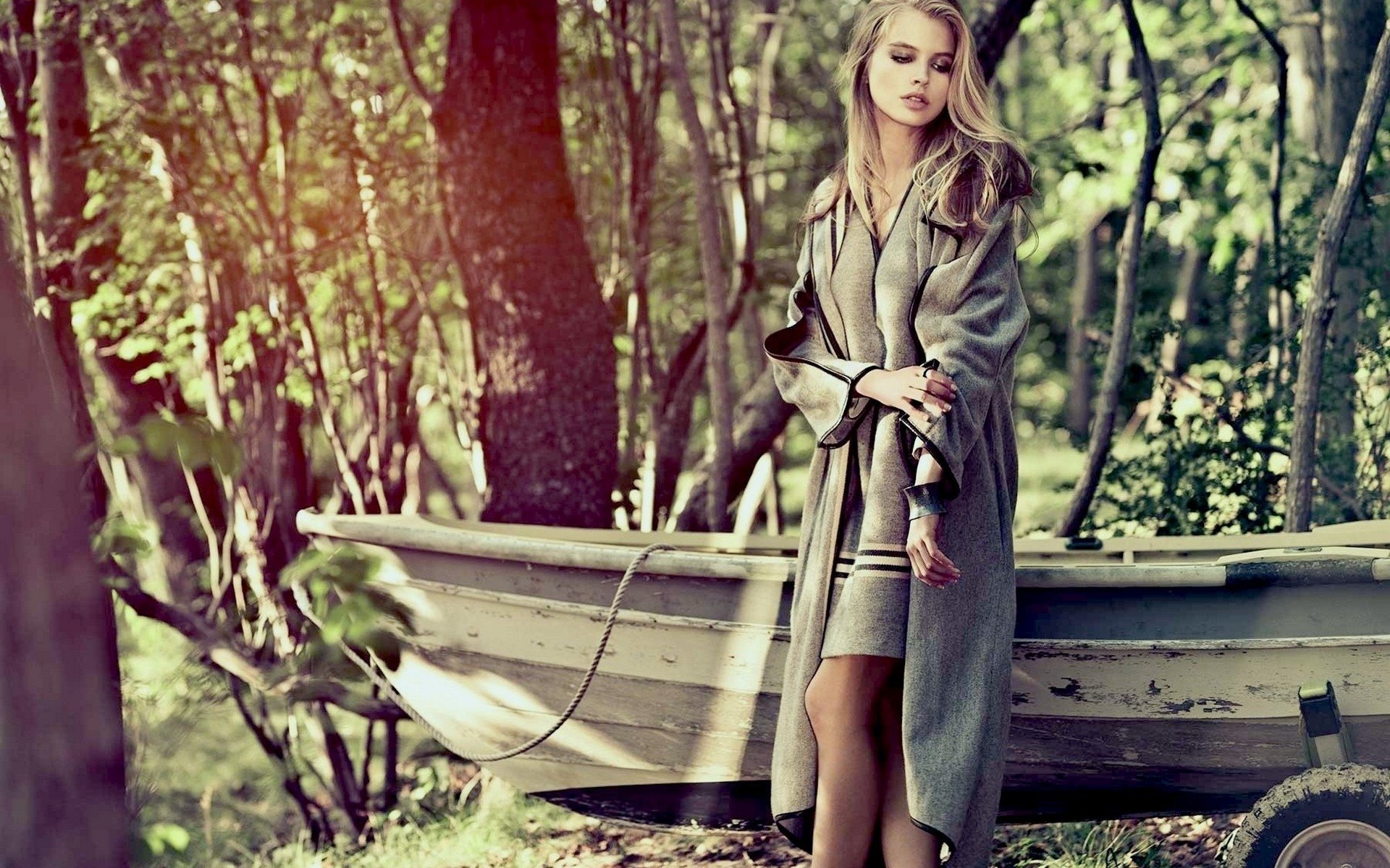 A serene image of Tanya Mityushina standing near an old boat in a forest, wearing a gray coat. The scene is bathed in warm sunlight, making it a captivating HD desktop wallpaper and background.