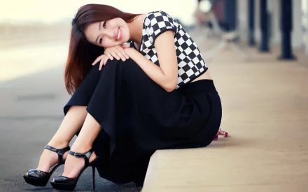 HD desktop wallpaper featuring an Asian woman sitting on a ledge, smiling, wearing a black and white checkered top, a black skirt, and high heels. The background includes a blurred urban setting.