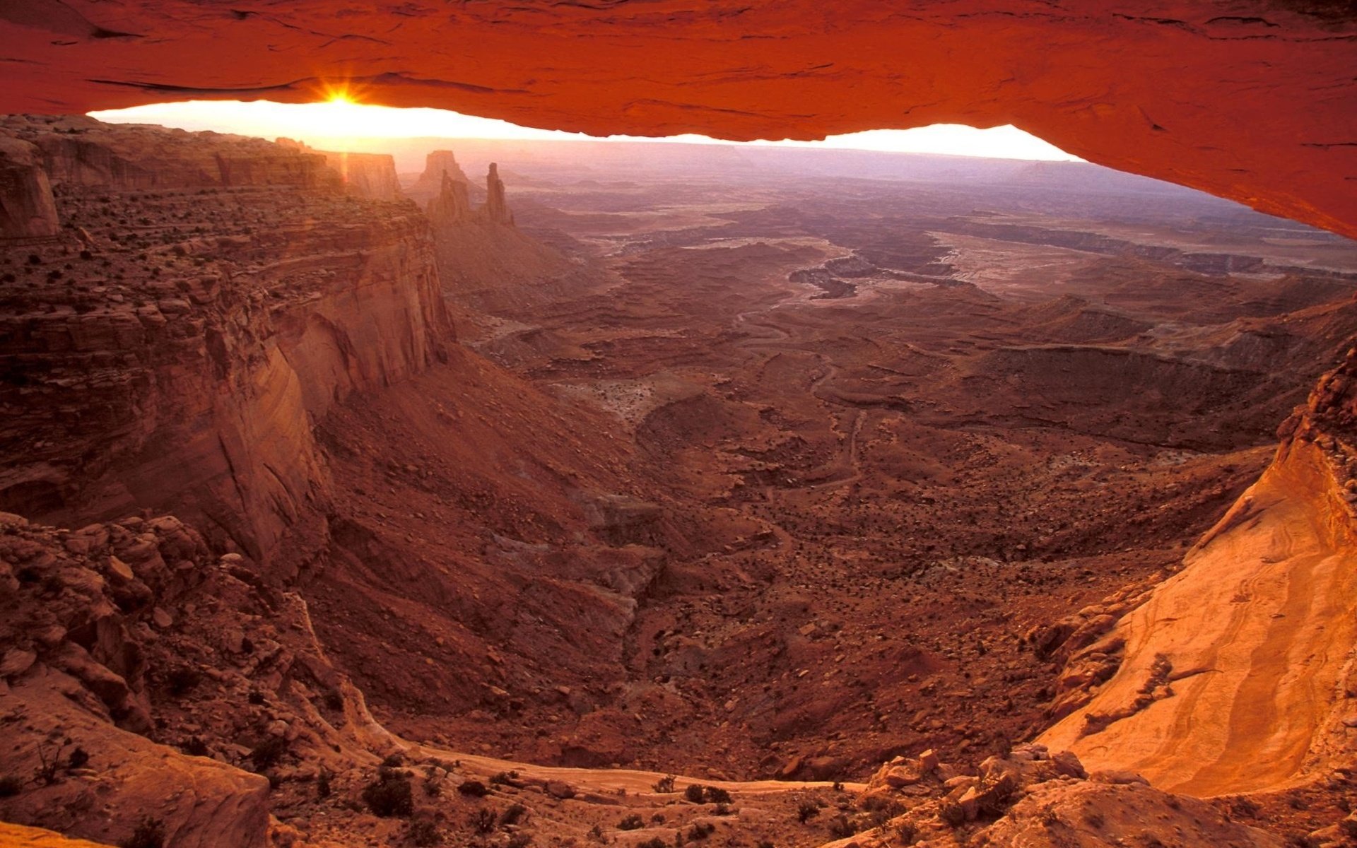 HD PC desktop wallpaper showcasing a majestic canyon landscape at sunrise, framed by a natural rock arch under warm, glowing hues.