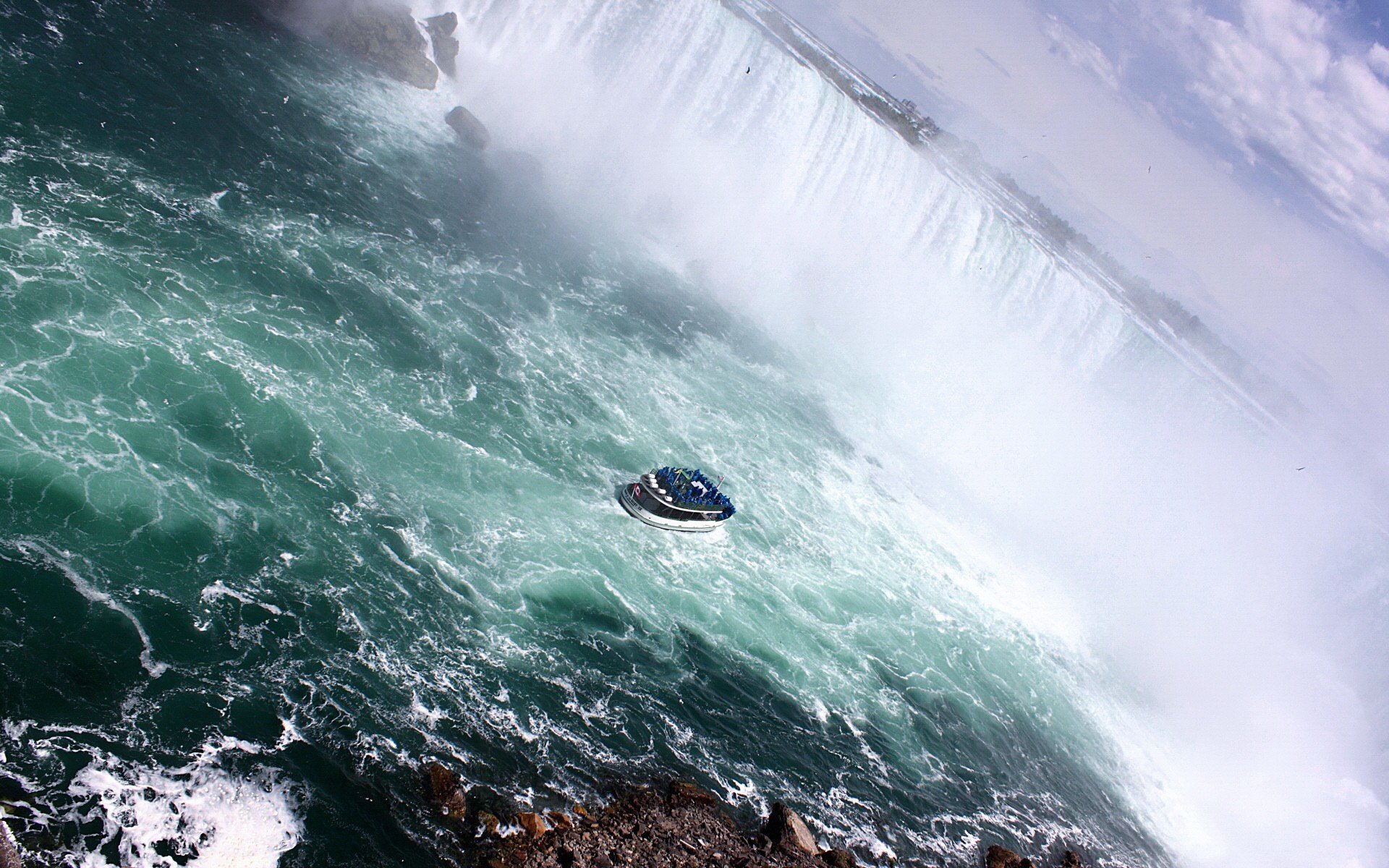 HD desktop wallpaper featuring the majestic Niagara Falls with a boat navigating through the turbulent waters and mist. The breathtaking natural scenery is enhanced by the vibrant blues and cascading water.