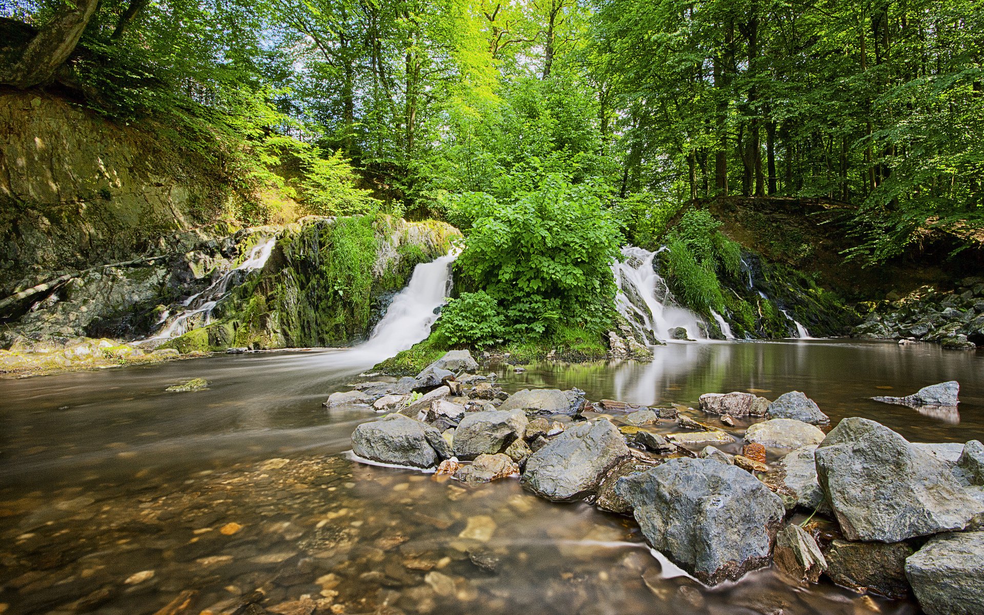 A serene waterfall cascades over rocks, surrounded by vibrant greenery, creating a tranquil natural setting. This HD image serves as a beautiful desktop wallpaper and background.
