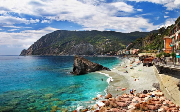 HD desktop wallpaper featuring the vibrant coastline and beach of Monterosso al Mare with colorful buildings and clear blue waters under a partly cloudy sky.
