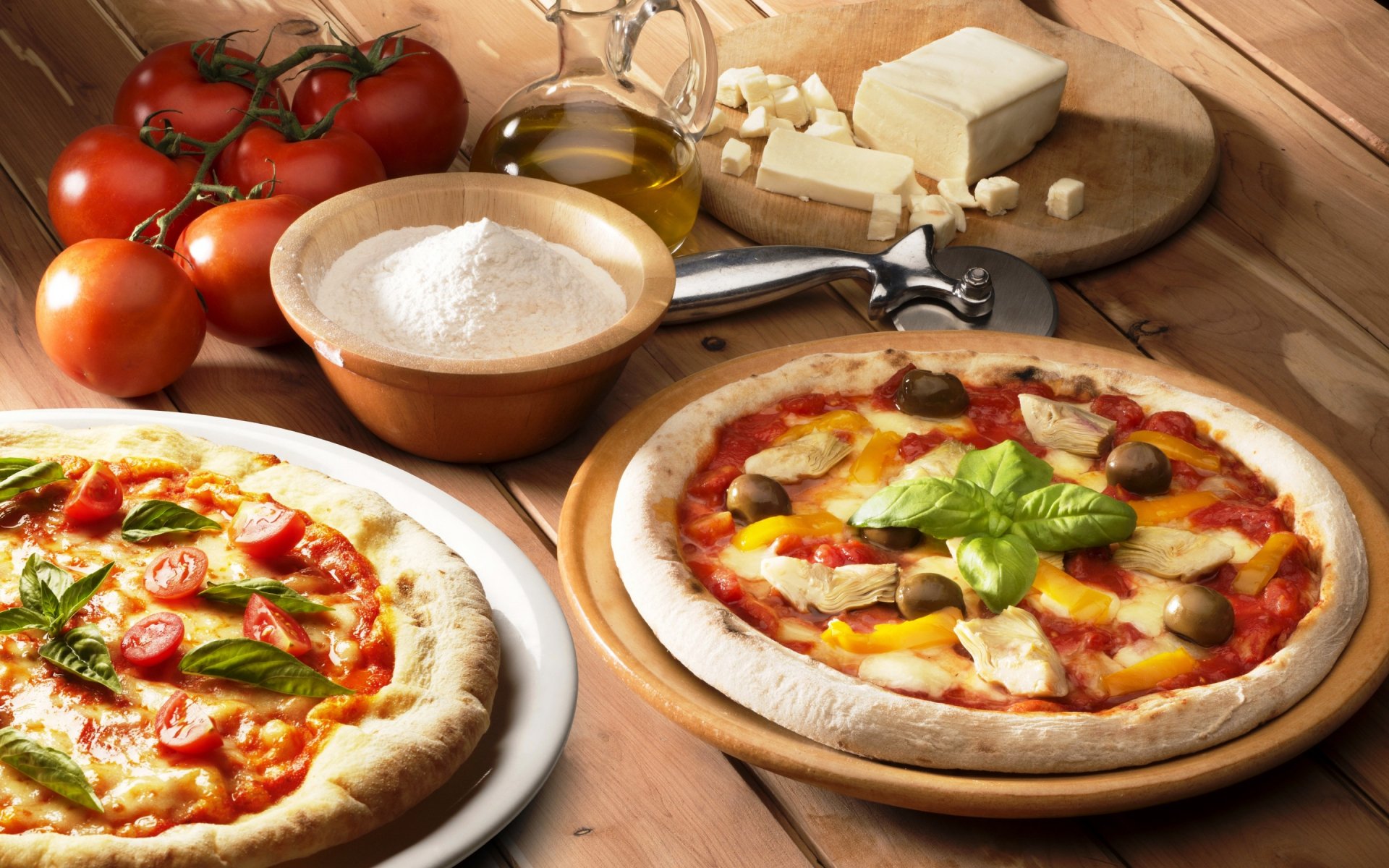 A vibrant assortment of homemade pizzas sits alongside fresh tomatoes, cheese, and flour, set against a wooden background, creating an appetizing HD wallpaper for food lovers.