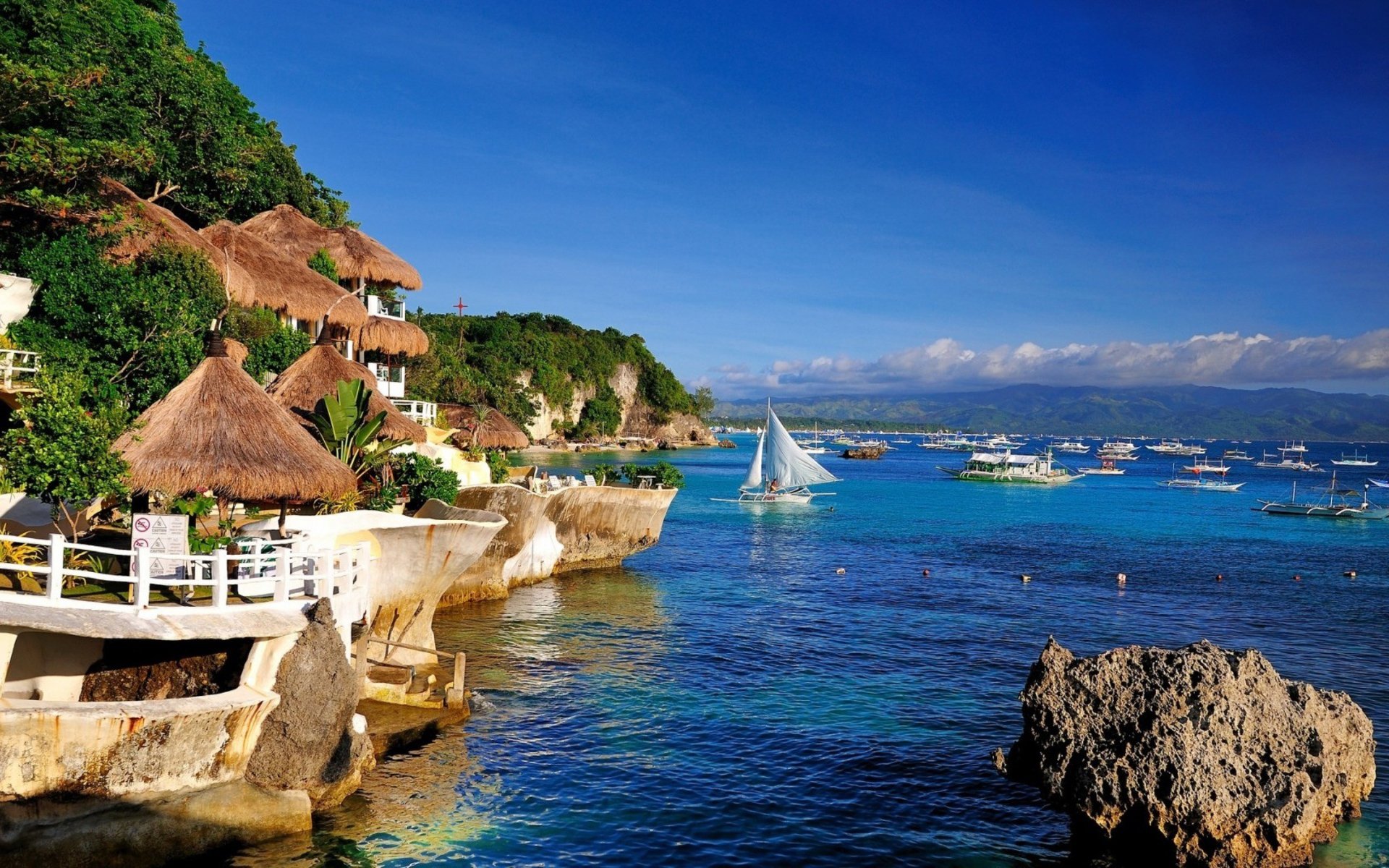 HD desktop wallpaper of a man-made resort with thatched-roof cottages along a rocky coastline and vibrant blue waters dotted with boats and a sailboat.