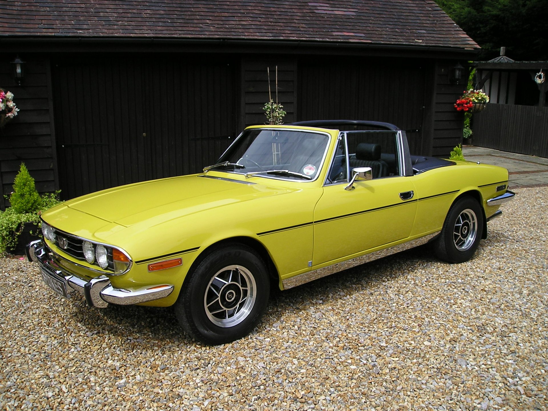 HD desktop wallpaper featuring a classic yellow Triumph Stag convertible parked on a gravel driveway with a rustic wooden building in the background.