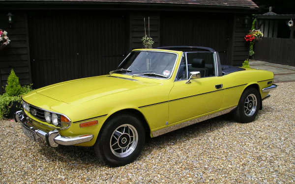 HD desktop wallpaper featuring a classic yellow Triumph Stag convertible parked on a gravel driveway with a rustic wooden building in the background.