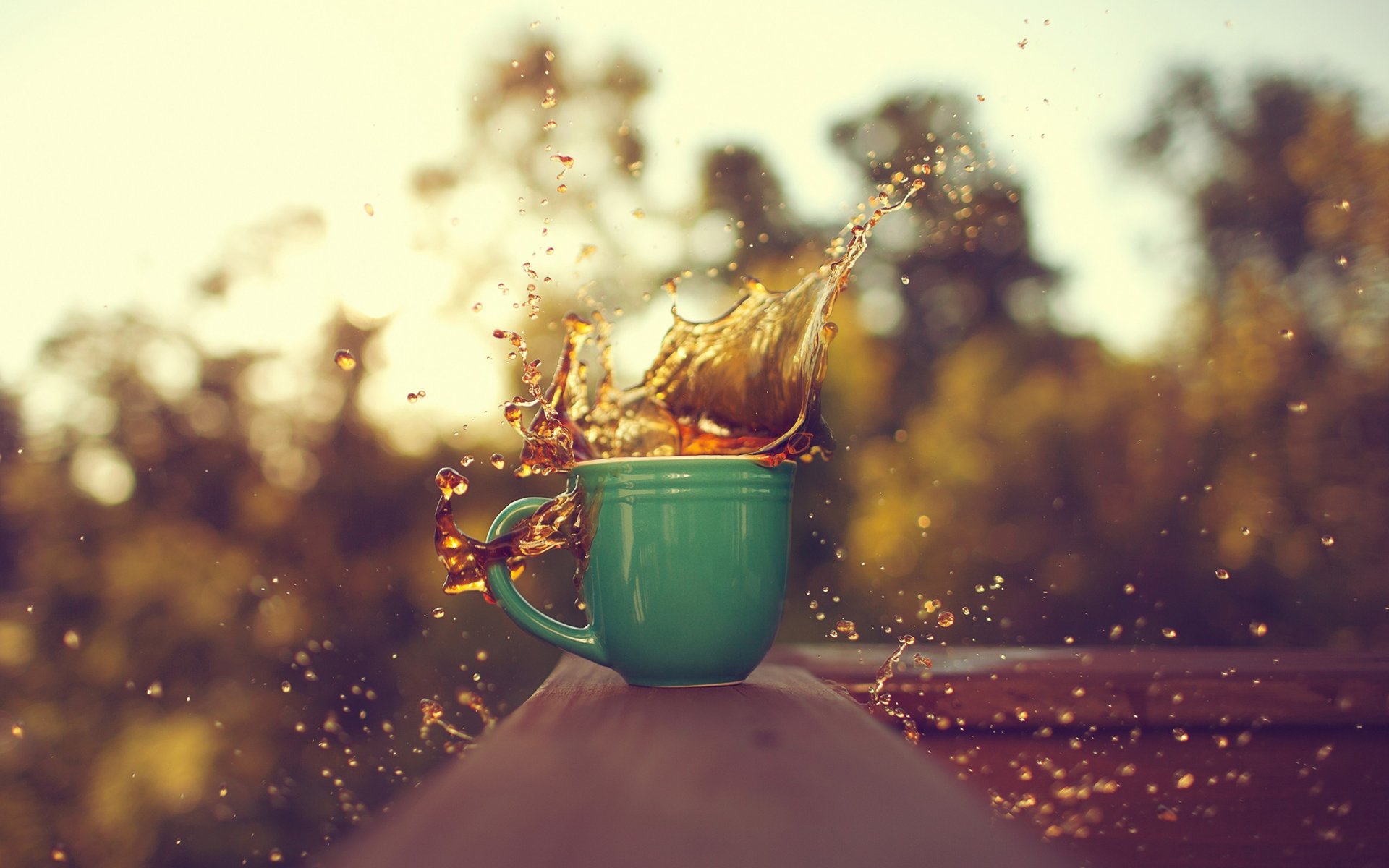 A vibrant HD PC desktop wallpaper showing coffee splashing out of a green mug against a softly blurred outdoor background.