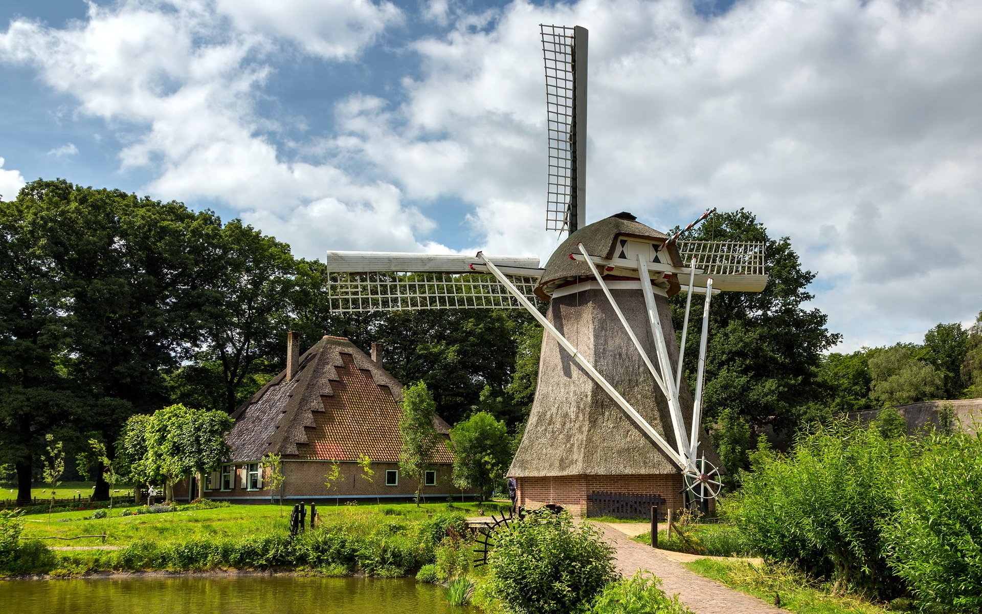HD PC desktop wallpaper showing a man-made windmill beside a thatched house, set in a lush green landscape under a partly cloudy sky.